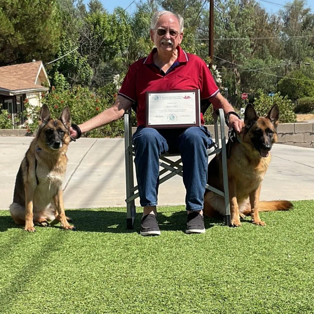 Ron Cable seated on a lawn holding his certificate, with a German Shepherd on each side